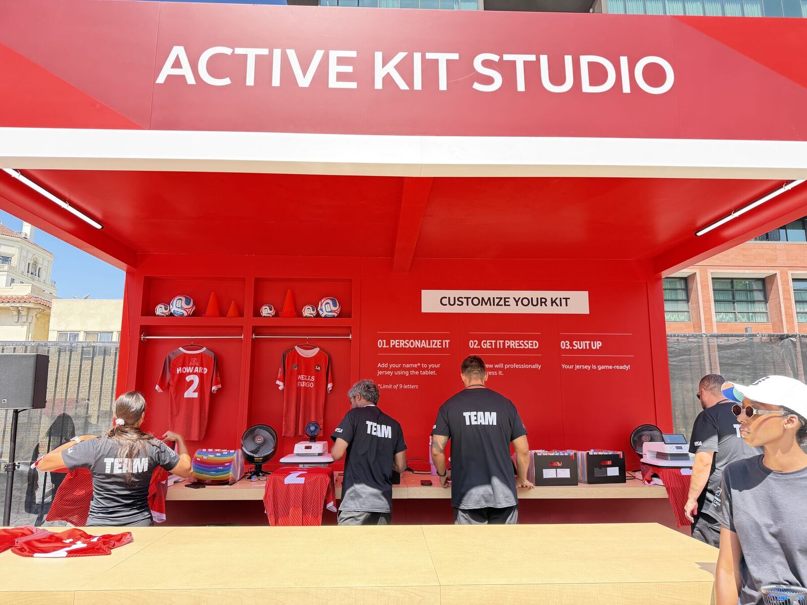 Red outdoor kit studio booth with locker bays holding personalized jerseys, tablet and press stations along the counter, and a crew in black TEAM shirts working the line