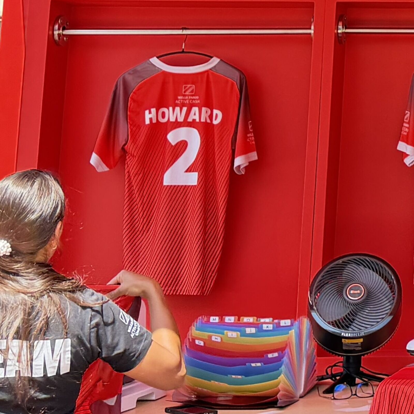Red jersey on a hanger in a red locker bay, pressed with the name HOWARD and the number 2 in white, while a crew member in a TEAM shirt works beside an alphabetized letter organizer